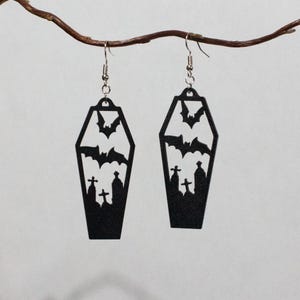 Könnte beinhalten: Ein Paar schwarze Ohrhänger in Sargform. Jeder Ohrring zeigt ein ausgeschnittenes Motiv einer Friedhofsszene mit Silhouetten von Grabsteinen und fliegenden Fledermäusen. Die Ohrringe hängen an silberfarbenen Metall-Ohrhaken.