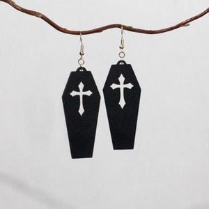 Könnte beinhalten: Ein Paar hängende Ohrringe in Form von schwarzen Särgen, die jeweils in der Mitte einen weißen Kreuz-Ausschnitt aufweisen. Die Ohrringe sind an silberfarbenen Metallhaken befestigt, die eine sichere Befestigung der Ohrschmuckstücke ermöglichen.