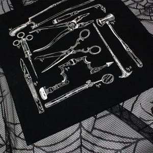 Schwarze Tragetasche mit einer weißen Illustration antiker chirurgischer Instrumente, darunter Sägen, Scheren und Spritzen. Das Design hat ein medizinisches Thema. Die Tasche ist aus dunklem Stoff.