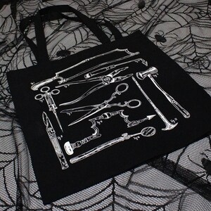 Schwarze Tragetasche mit einer weißen Illustration verschiedener chirurgischer Instrumente. Die Werkzeuge umfassen Sägen, Scheren und andere medizinische Instrumente. Die Tasche hat schwarze Griffe und besteht aus robustem Material.