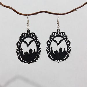 Könnte beinhalten: Ein Paar schwarze Ohrhänger mit einem ovalen Rahmen und einer filigranen Bordüre. In der Mitte jedes Ohrrings befindet sich die Silhouette eines Friedhofs mit Grabsteinen und einer fliegenden Fledermaus, befestigt an silberfarbenen Metallhaken.