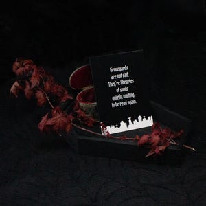 Eine schwarze Sarg-förmige Box mit rotem Samt-Innenraum, die eine Karte mit dem Text "Graveyards are not sad. They're libraries of souls quietly waiting to be read again." enthält. Getrocknete rote Blätter und eine kleine dekorative Box dienen als Dekoration.