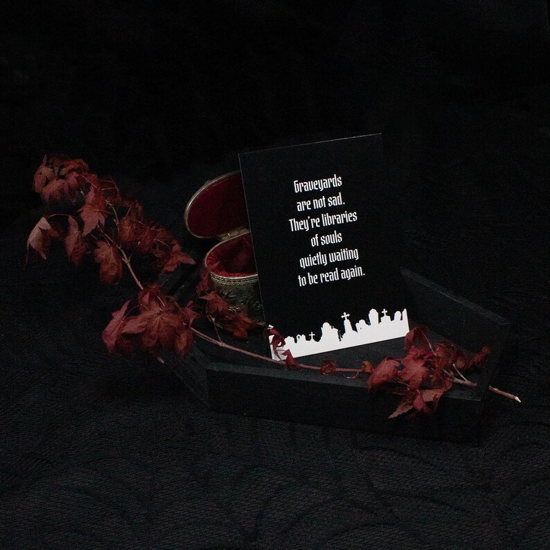 Eine schwarze Sarg-förmige Box mit rotem Samt-Innenraum, die eine Karte mit dem Text "Graveyards are not sad. They're libraries of souls quietly waiting to be read again." enthält. Getrocknete rote Blätter und eine kleine dekorative Box dienen als Dekoration.