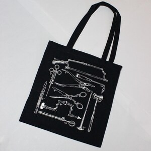 Schwarze Tragetasche mit einer weißen Illustration von chirurgischen Werkzeugen im Vintage-Stil. Das Design umfasst Sägen, Scheren und andere medizinische Instrumente. Die Tasche hat zwei schwarze Griffe.