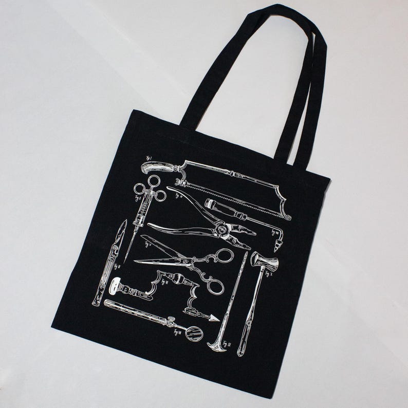 Schwarze Tragetasche mit einer weißen Illustration von chirurgischen Werkzeugen im Vintage-Stil. Das Design umfasst Sägen, Scheren und andere medizinische Instrumente. Die Tasche hat zwei schwarze Griffe.