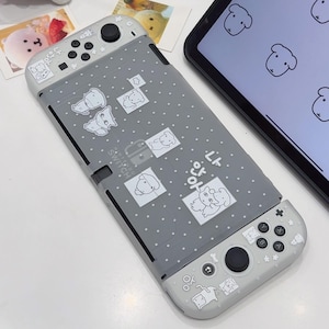 Op de afbeelding: Een grijze Nintendo Switch console met een beschermhoes voorzien van een wit stippenpatroon en diverse cartoon-dierillustraties. De hoes bevat Koreaanse teksttekens en kleine sterpictogrammen op de behuizing en beide controllers.
