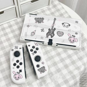 Puede incluir: Una consola de juegos blanca y dos mandos a juego con un estampado gráfico en negro y rosa. El diseño incluye una guitarra, notas musicales, un corazón, una jeringuilla, huesos y personajes de dibujos animados con cuernos o halos.