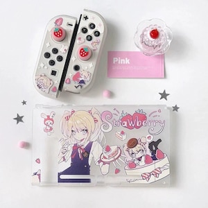 May include: A clear protective case and controller covers for a gaming console, featuring anime-style illustrations of a blonde character, strawberries, and cake. Includes a small pink card with the text Pink and a clear, round decorative topper.