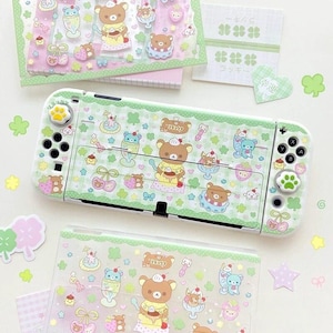 Könnte beinhalten: Eine weiße Spielkonsole mit einem hellgrünen Karomuster-Skin, verziert mit Rilakkuma-Bären, Desserts und Kirschen. Die Konsole enthält passende grüne Joystick-Kappen in Pfotenform und eine Schutzhülle mit dem gleichen Muster.