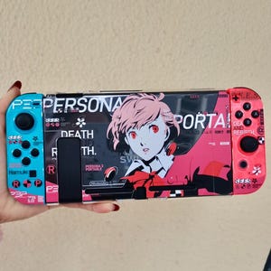 May include: A handheld gaming console featuring a custom skin with Persona 3 Portable graphics. The left controller is light blue, the right is red, and the center console displays a character with pink hair, the text Persona, and Death and Rebirth.