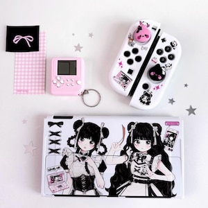 Puede incluir: Una base de carga blanca para consola de juegos y dos mandos decorados con ilustraciones de personajes estilo anime en negro y rosa, una pequeña consola portátil rosa con llavero y una tarjeta de cuadros vichy rosa con un adhesivo de lazo negro.