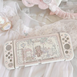 Puede incluir: Una funda protectora blanca para consola de juegos con una ilustración detallada de un oso de peluche, un conejo y un perro pequeño en un jardín con fresas y flores. Incluye cubiertas laterales a juego con pequeños motivos florales y animales.