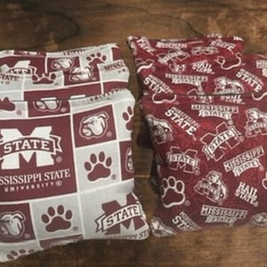 May include: Set of four Mississippi State University cornhole bags. Two bags feature a maroon and white pattern with the university logo, paw prints, and text. The other two bags have a maroon pattern with the university logo and text.