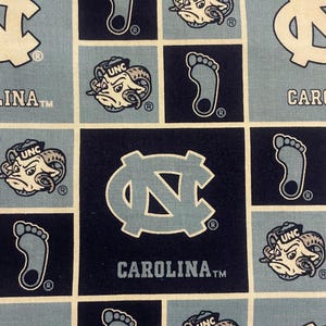 May include: A repeating fabric pattern in a grid of light blue and navy blue squares. Each square features a white and light blue design of a foot, a ram head with "UNC" on its head, and the letters "UNC" in a stylized font. The text "CAROLINA" is also present.