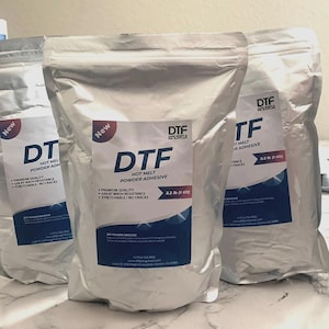 Könnte beinhalten: Drei silberne Beutel mit DTF-Heißschmelz-Pulverkleber. Jeder Beutel hat ein blau-weißes Etikett mit dem Text "DTF" und "1 KG". Die Beutel sind mit "Neu" gekennzeichnet und listen Eigenschaften wie Waschbeständigkeit auf.