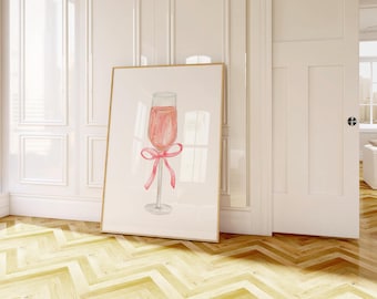 May include: A framed watercolour print of a champagne flute filled with pink liquid and a pink bow. The artwork has a cream background and a gold frame. The print is displayed in a room with white walls and a wooden floor.
