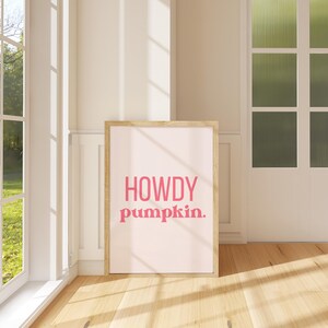 Può includere: Una stampa incorniciata con le parole "HOWDY pumpkin." in lettere rosa su sfondo rosa chiaro. La stampa è in una cornice di legno chiaro ed è appoggiata a un muro in una stanza con una finestra e pavimento in legno.