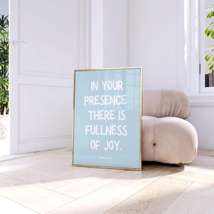 May include: A framed art print with a gold border, featuring the text "IN YOUR PRESENCE THERE IS FULLNESS OF JOY." The background of the print is a light blue color. The print is displayed in a bright room with a beige armchair and a window.