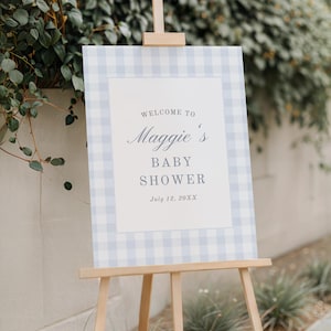 May include: A baby shower welcome sign on a wooden easel. The sign features a light blue and white gingham border and a white centre with the text "Welcome to Maggie's Baby Shower" and the date July 12, 20XX.