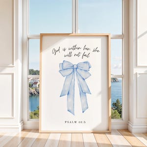Puede incluir: Impresión enmarcada con un marco marrón claro. La imagen presenta un lazo azul claro en acuarela sobre un fondo blanco. El texto "God is within her, she will not fail" está encima del lazo, con "PSALM 46:5" debajo.
