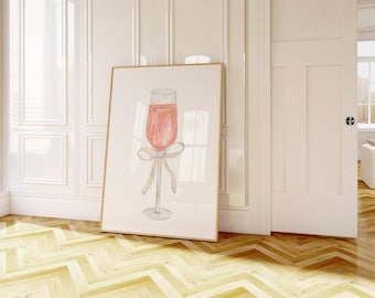 May include: A framed watercolour print depicting a champagne glass filled with a pink drink. The glass has a silver stem and a bow tied around the base. The artwork is displayed in a room with white walls and a wooden floor.