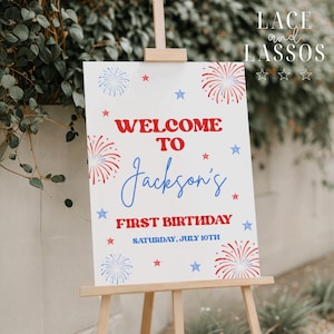 May include: A white rectangular birthday welcome sign on a wooden easel featuring red and blue firework illustrations and stars. The text reads: WELCOME TO Jackson's FIRST BIRTHDAY SATURDAY, JULY 10TH. The design uses a patriotic red and blue color scheme.