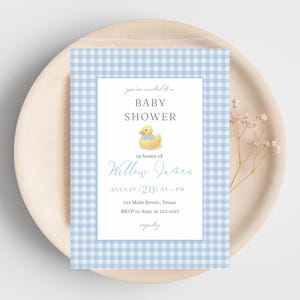 May include: Baby shower invitation with a blue gingham border. The invitation features the text "Baby Shower" and "Willow James" with a watercolour illustration of a yellow rubber ducky wearing a blue bow tie. The invitation is on a wooden plate.