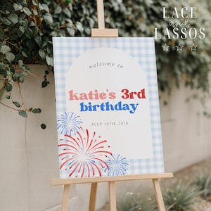 May include: A wooden easel displays a sign with a blue gingham border. The sign reads "welcome to katie's 3rd birthday" with red and blue text, and features firework illustrations. The date "JULY 10TH, 2026" is also displayed.
