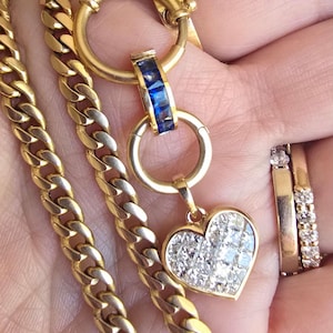 Op de afbeelding: Een gouden schakelketting met een ronde sluiting, een cilindervormige bedel met blauwe vierkante stenen en een hartvormige hanger bezet met heldere vierkante stenen. Twee gouden ringen met heldere stenen worden aan de vinger gedragen.