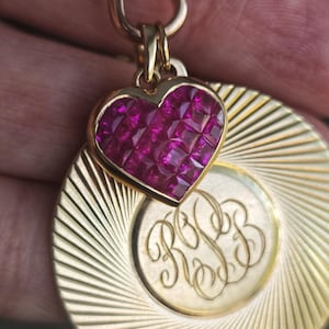 May include: A gold-toned pendant featuring a heart-shaped charm filled with pink gemstones. Below the heart is a circular pendant with engraved initials and a sunburst design.