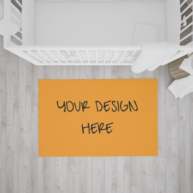 以下が含まれることがあります： 黒い文字で「YOUR DESIGN HERE」と書かれたオレンジ色の長方形のラグ。 ラグは明るい木製の床にあり、背景には白いベビーベッドと寝具があります。