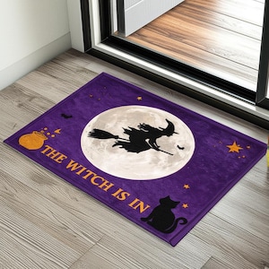 Könnte beinhalten: Lila Halloween-Fußmatte mit Vollmond, Hexe auf einem Besen und dem Text "THE WITCH IS IN". Mit schwarzer Katze, Sternen und einem Kessel. Eine festliche Willkommensmatte für Halloween.