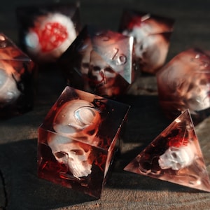 Peut inclure: Un ensemble de dés en résine avec un motif de crâne. Les dés, de formes variées, dont des cubes et des pyramides, présentent des accents rouges et blancs. Ils sont présentés sur une surface en bois.