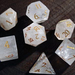 May include: A collection of seven clear, multi-sided dice with gold-colored numbering. The dice are arranged on a dark, textured surface. The dice are of various shapes, including a four-sided pyramid, a six-sided cube, and an eight-sided die.