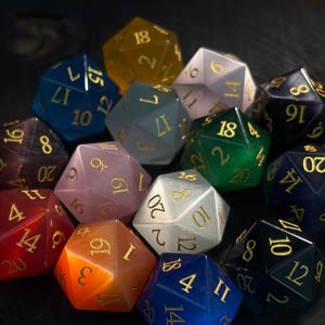 May include: A collection of polyhedral dice in various colors, including blue, orange, and green. Each die has gold-colored numbers. The dice are arranged on a dark surface, creating a visually appealing display for tabletop gaming.