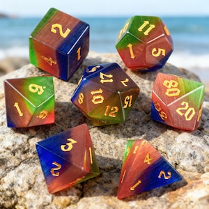 May include: Seven multi-sided dice in different shapes, each with a colour gradient of red, green, and blue. The dice have gold-coloured numbers. They are arranged on a rock surface, with the sea in the background.
