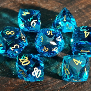 May include: Seven blue, faceted dice with gold numerals are displayed. The dice are in various shapes, including cubes and pyramids. The dice are arranged on a dark, textured surface. The dice are for role-playing games.