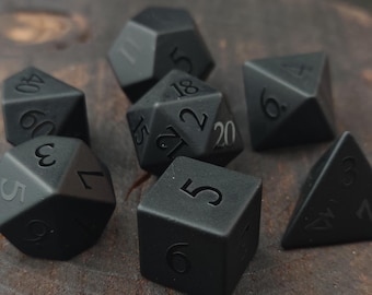 May include: A collection of matte black polyhedral dice, including D4, D6, D8, D10, D12, and D20. Each die features white numbering. The dice are arranged on a dark, textured surface, suitable for tabletop gaming.