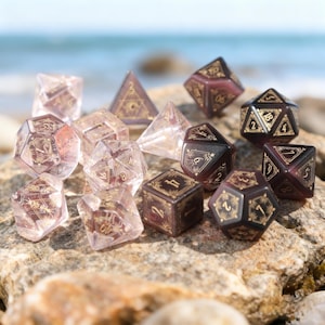 May include: A collection of polyhedral dice in various shapes and sizes, including D4, D6, D8, D10, D12, and D20. The dice are clear and burgundy with gold-colored numbering and decorative accents. The dice are arranged on a rock with the ocean in the background.