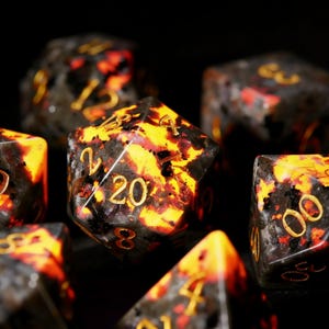 May include: Close-up of several polyhedral dice with a black and orange speckled design. The numbers are gold. The dice are for tabletop games.