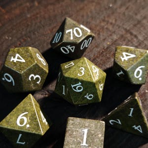 Puede incluir: Un conjunto de siete dados verdes con números blancos, incluyendo un dado de cuatro caras, uno de seis, ocho, diez, doce, veinte caras y un dado de porcentaje. Los dados están hechos de un material moteado similar a la piedra, ideal para juegos.