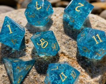 May include: A set of seven translucent blue dice with gold numbering, resting on a rock. The dice are various shapes, including a d4, d6, d8, d10, d12, d20, and a percentile die. The dice are for tabletop games.