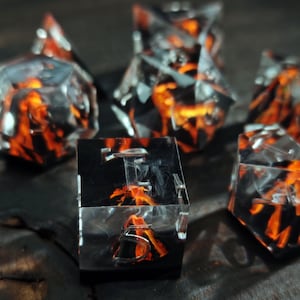 Puede incluir: Un conjunto de dados poliédricos con un diseño de fuego. Los dados son transparentes con llamas negras y naranjas en el interior, creando un efecto visual único. Los dados están dispuestos sobre una superficie oscura.