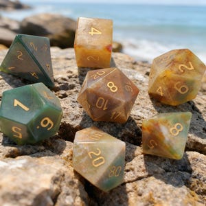 May include: A collection of seven polyhedral dice in various shapes, resting on a rock near the ocean. The dice are translucent with earthy tones of green, brown, and yellow, and feature gold numbering. Ideal for tabletop games.