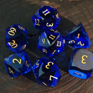 Può includere: Set di sette dadi poliedrici blu con numeri dorati. I dadi sono sfaccettati e sembrano essere fatti di un materiale traslucido. Sono disposti su una superficie scura e strutturata, adatti per giochi di ruolo.
