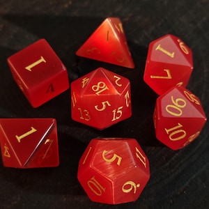 May include: A set of seven polyhedral dice in a translucent red finish with a cat's eye effect. Each die features engraved numbers filled with gold-colored ink, including a d20, d12, d10, d8, d6, d4, and a percentile die with tens digits.