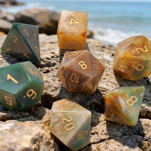 May include: A collection of gemstone dice in various shapes and colors, including green, brown, and yellow. The dice are set against a rocky surface with the ocean in the background. Each die features gold-colored numbers.