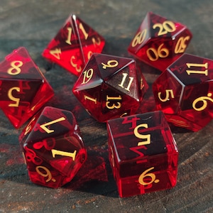 May include: A set of seven translucent red polyhedral dice with gold-coloured numbers. The collection includes a d20, d12, d10, d8, d6, d4, and a percentile die, all featuring sharp edges and clear, legible numerals etched into the deep red resin surfaces.