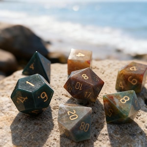May include: A collection of gemstone dice in various shapes and colors, including green, brown, and amber. The dice are resting on a rock with a blurred ocean background. The numbers are in gold.