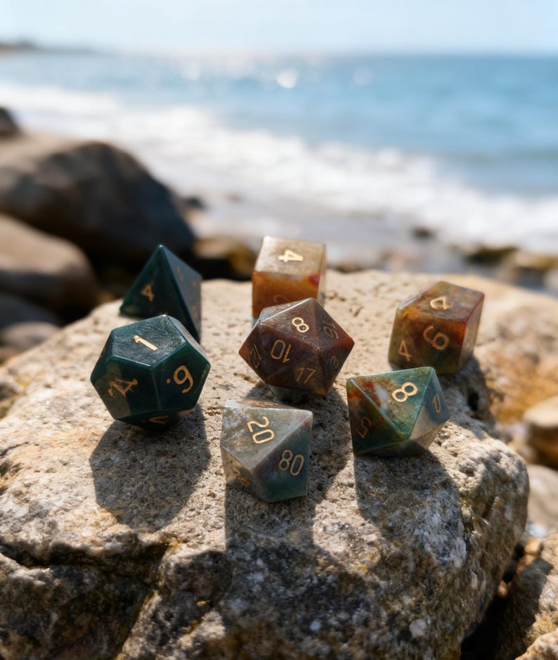 May include: A collection of polyhedral dice in various colors, including green, brown, and clear, with gold numbering. The dice are resting on a rock with the ocean in the background. Ideal for tabletop gaming.
